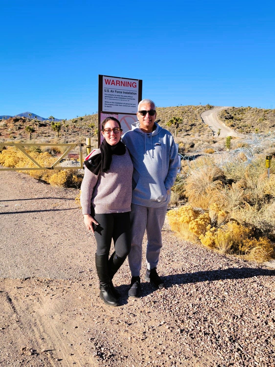 Tourists in a rugged, desert landscape with sparse vegetation, gazing towards the distant, heavily fenced perimeter of Area 51 under a vast, blue sky, 